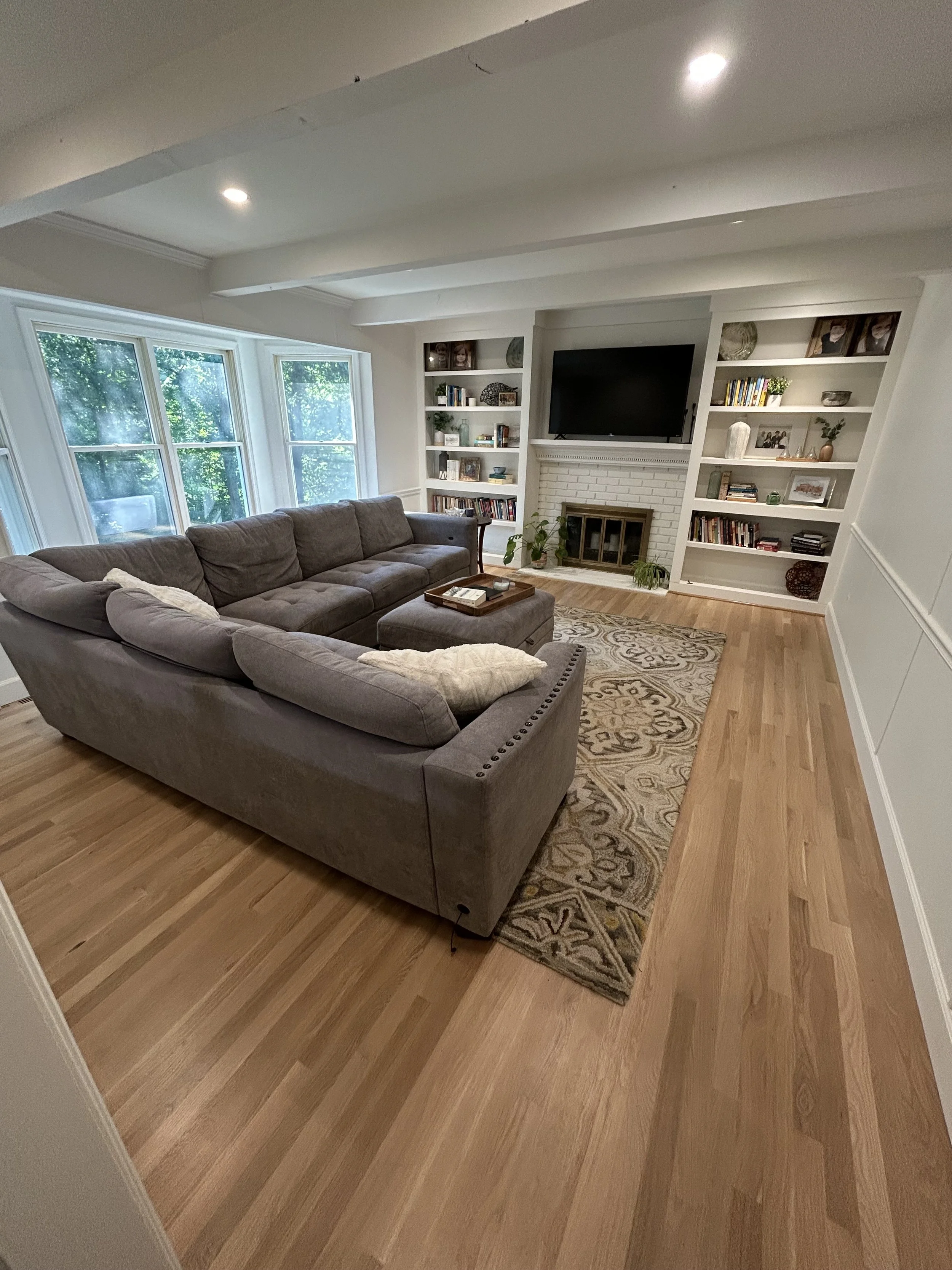 Living room with a gray sectional sofa, a patterned area rug, built-in white shelves with books and decorative items, a white brick fireplace, a large TV above the fireplace, and three windows allowing natural light to enter.