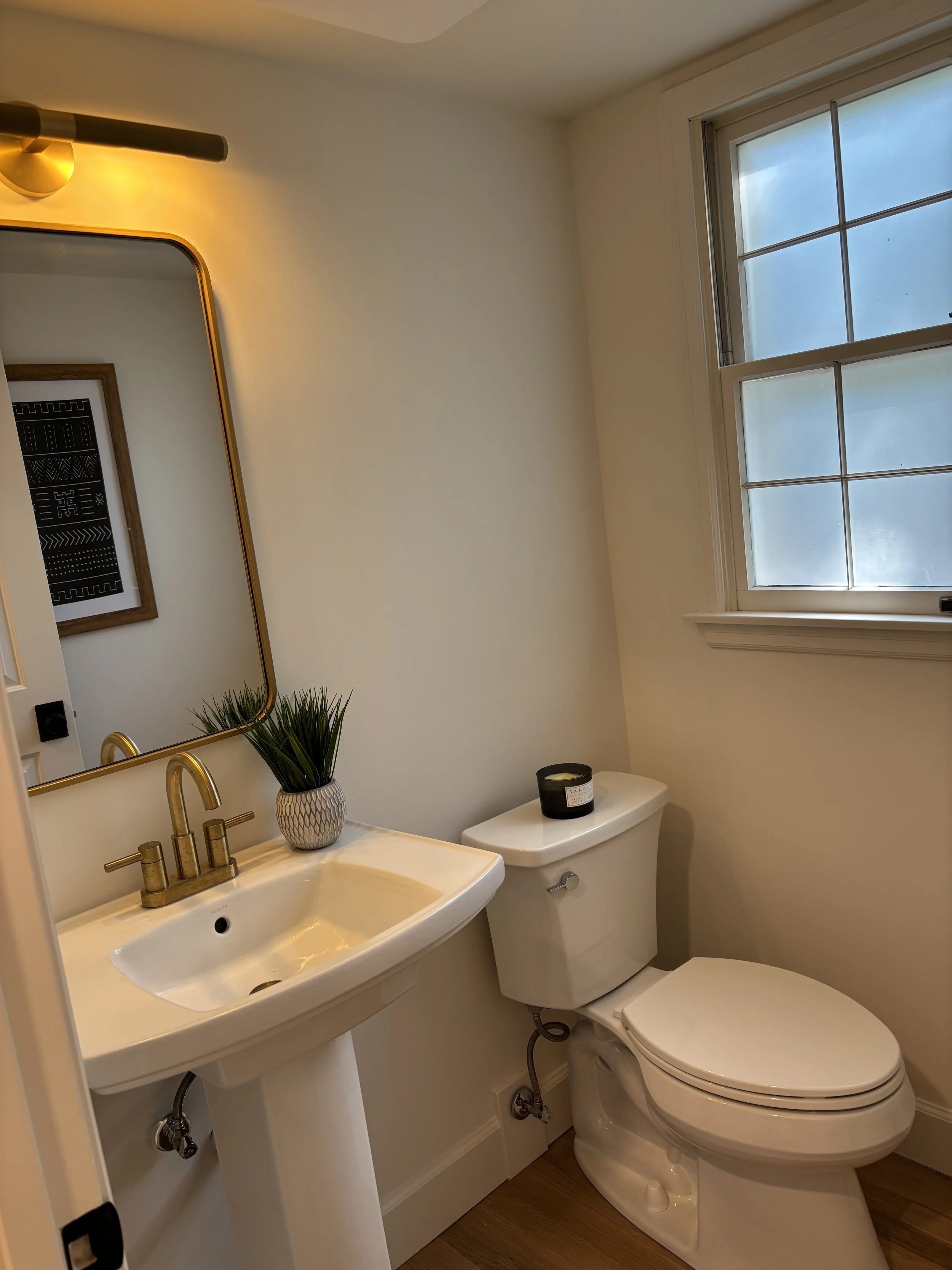Small bathroom with white sink, gold faucet, mirror, potted plant, and toilet with a candle on top. There is a window with frosted glass.