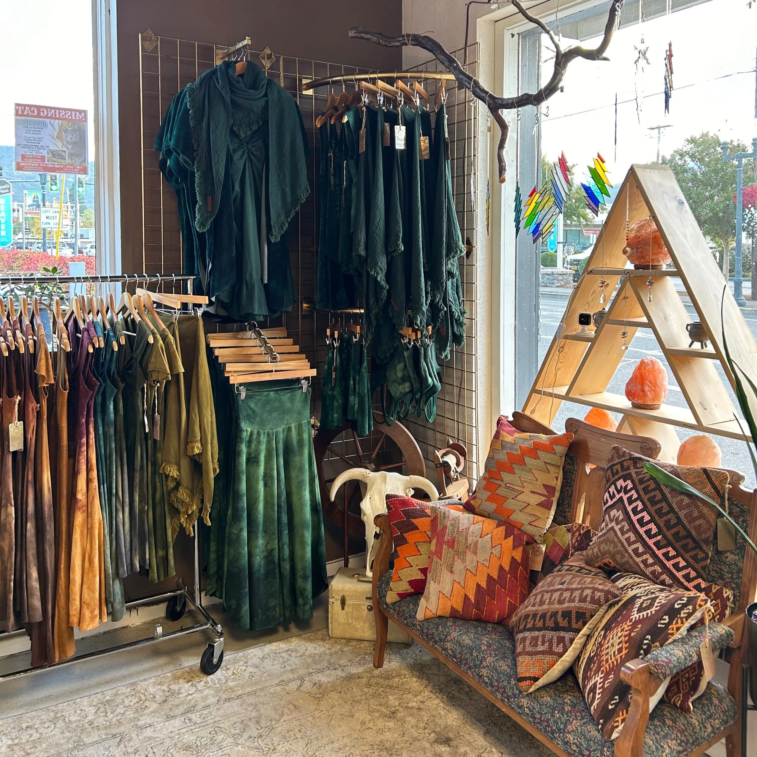 A display of green and multicolored clothing, including skirts, tops, and scarves, in a boutique with a sofa adorned with patterned pillows, a wooden triangular shelf with Himalayan salt lamps, and a window showing a street view.