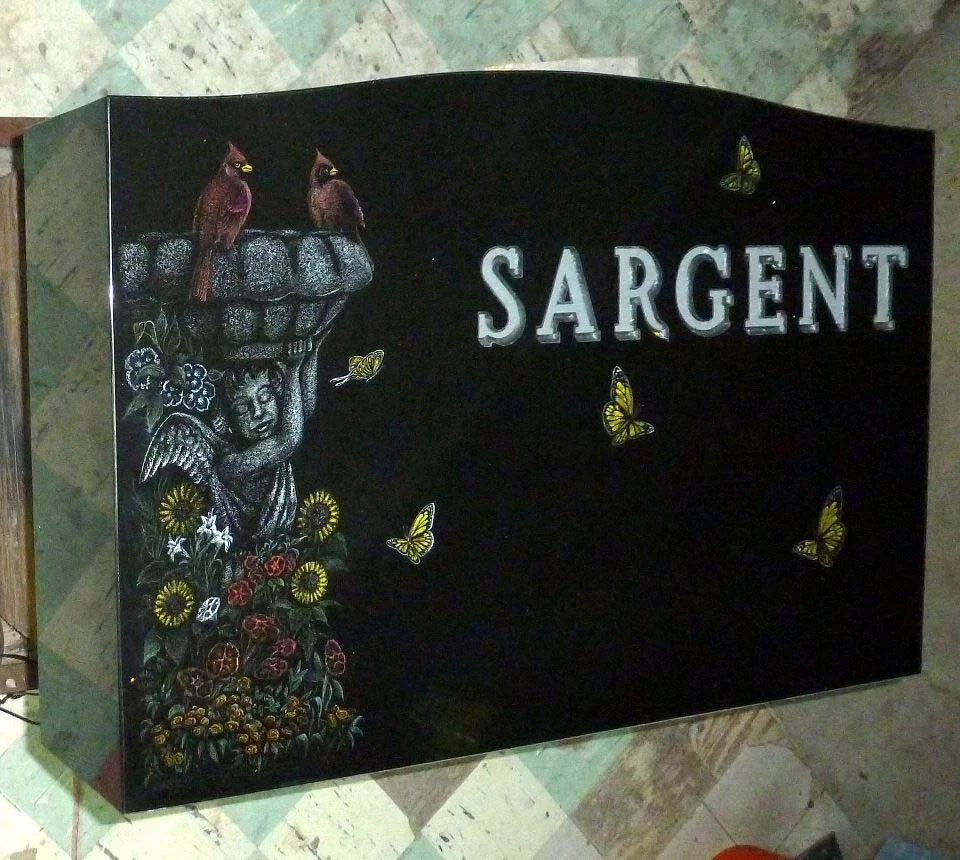 Headstone with the name 'Sargent' featuring carved cardinals, an angel, flowers, and butterflies on a dark stone background.