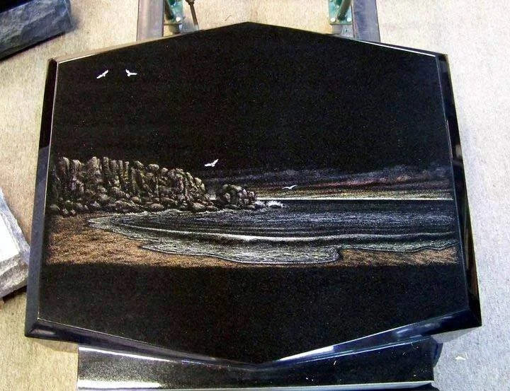 Engraved stone artwork depicting a coastal landscape with cliffs, waves, and seagulls on a dark background.