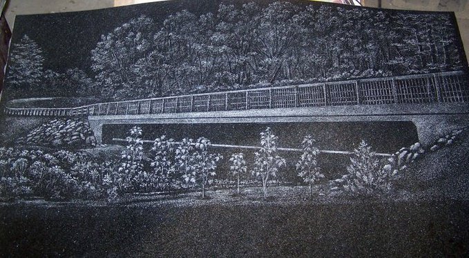 Etched artwork of a landscape on a dark surface, featuring a bridge surrounded by trees and vegetation.