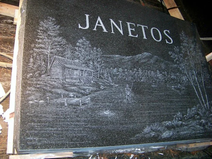Black granite engraving of a rural scene with a log cabin, trees, a fisherman, water, and the name "Janetos" at the top.