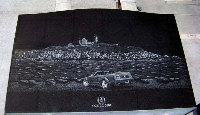 Engraved stone with a scenic view of a lighthouse on a rocky coastline and a convertible car in the foreground. The date "Oct. 30, 2004" is inscribed at the bottom.