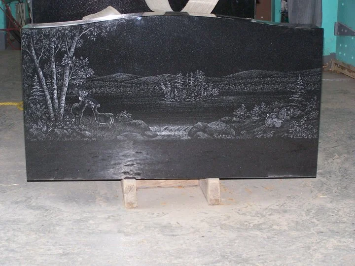 A polished black granite tombstone featuring an etched landscape scene with trees, a deer, and a natural setting.