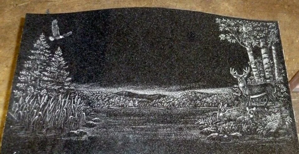 Engraved granite slab depicting a nature scene with a lake, trees, mountains, an eagle in flight, and deer standing beside a forest.