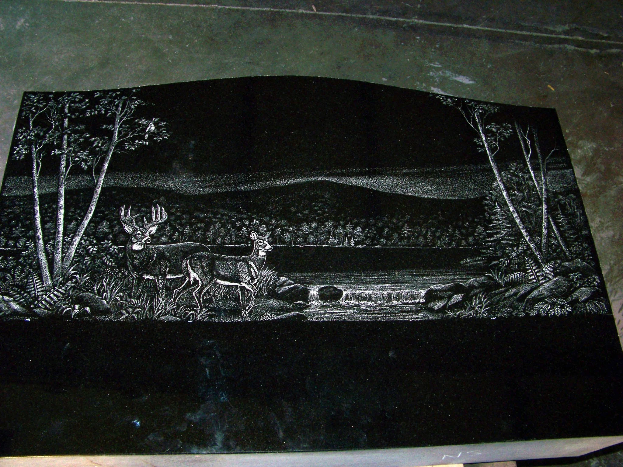 Engraved black stone with forest scene; features two deer, trees, and a stream.
