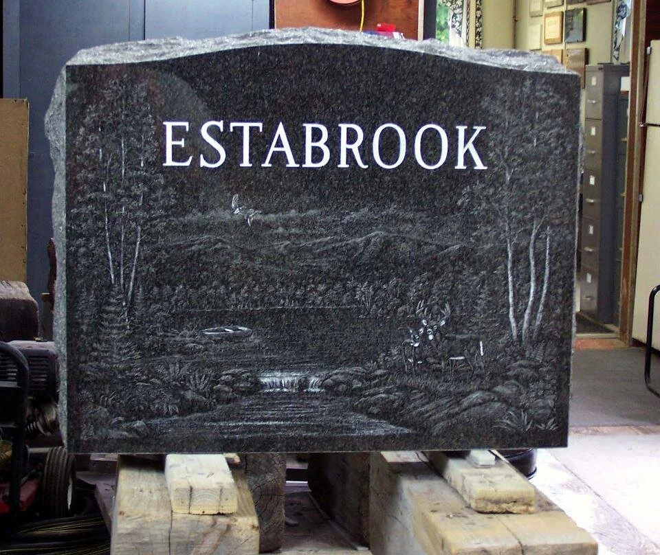Granite tombstone with the name 'Estabrook' and a forest landscape engraving, placed on wooden supports.