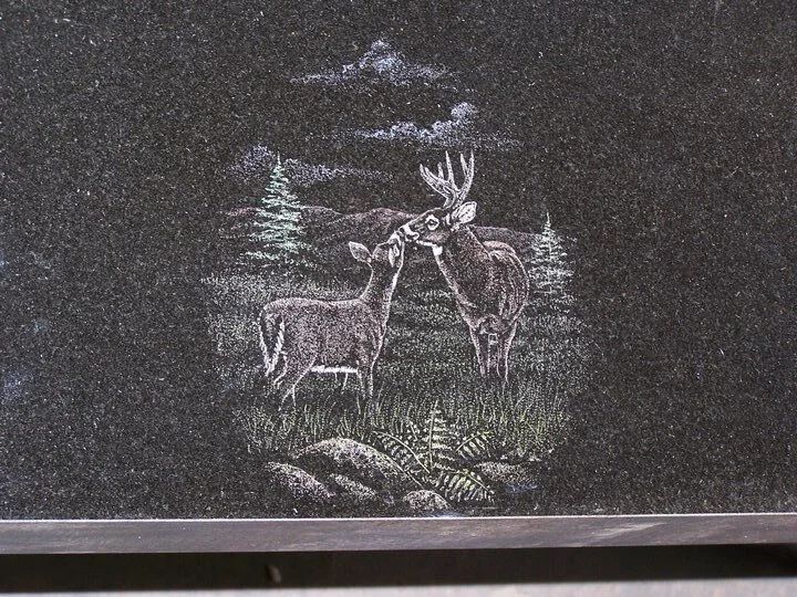 Etching of two deer in a forest scene on a black stone surface.