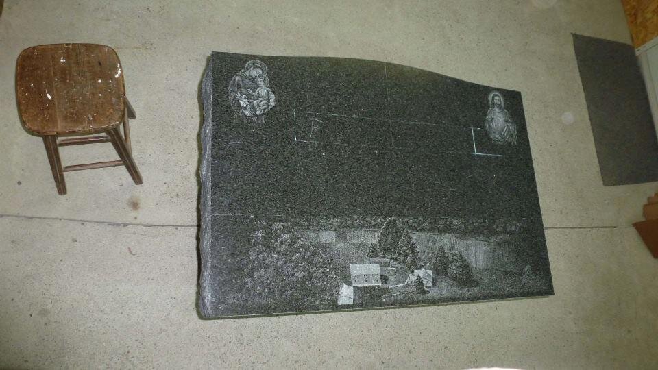 A large, dark granite tombstone featuring engraved images of religious figures and a landscape scene, placed next to a wooden stool on a concrete floor indoors.