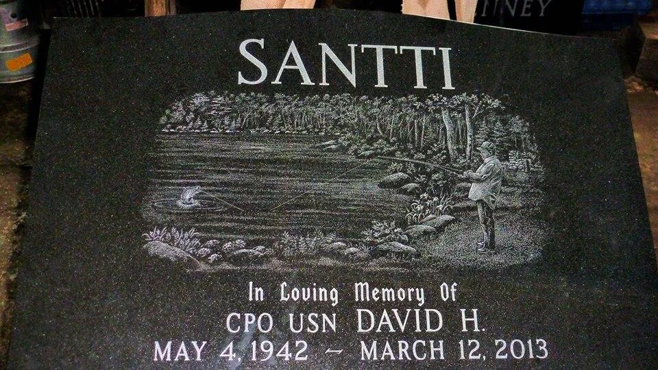 Headstone with engraving for 'Santti'. It features an illustration of a person fishing in a scenic landscape. The text reads 'In Loving Memory Of CPO USN David H. May 4, 1942 – March 12, 2013.'