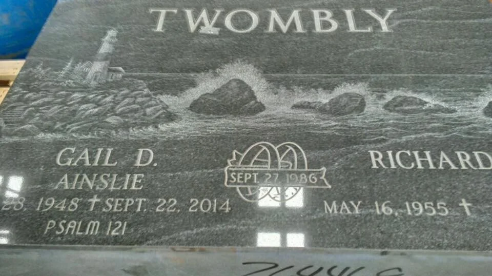 Engraved granite headstone with the name "Twombly," featuring lighthouse and ocean waves design with dates and names: Gail D. Ainslie, born May 28, 1948, died Sept. 22, 2014, and Richard, born May 16, 1955. Includes a ring symbol dated Sept. 27, 1986