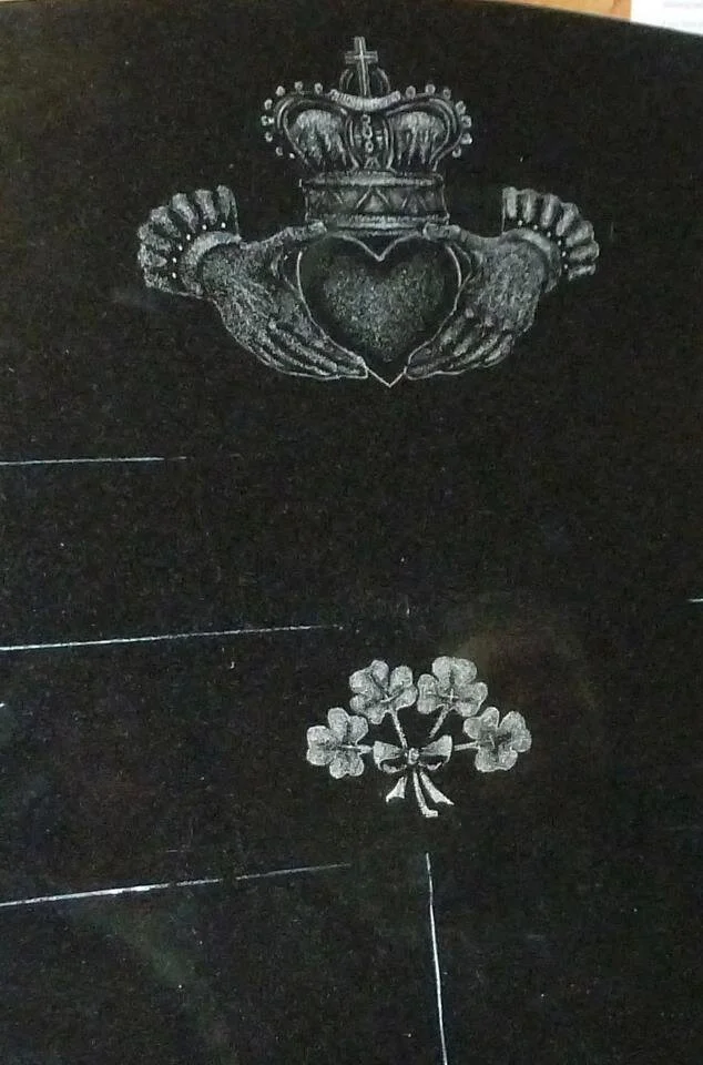 Etched design of Claddagh symbol with hands holding a heart topped by a crown, and below it a cluster of shamrocks tied with a ribbon on a dark surface.