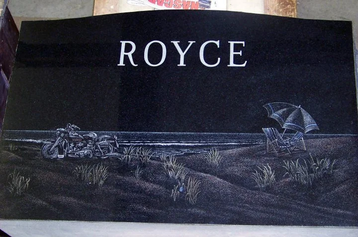Engraved tombstone with the name 'Royce' featuring a beach scene with a motorcycle, beach chairs, and an umbrella.