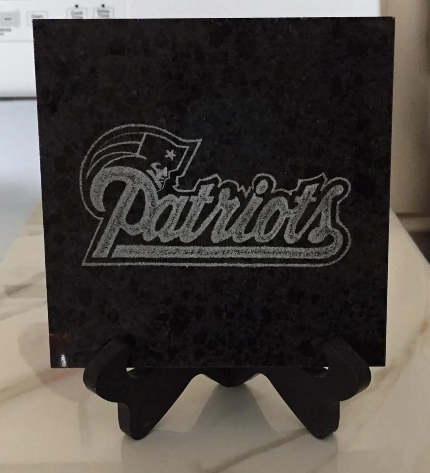 Dark stone tile with "Patriots" and a helmeted figure logo etched on it, placed on a wooden stand.