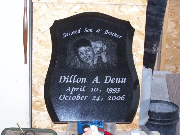 Black granite tombstone for Dillon A. Denu, labeled 'Beloved Son & Brother', with birth and death dates April 10, 1993, October 24, 2006. Includes etched portrait of a young person with a dog.