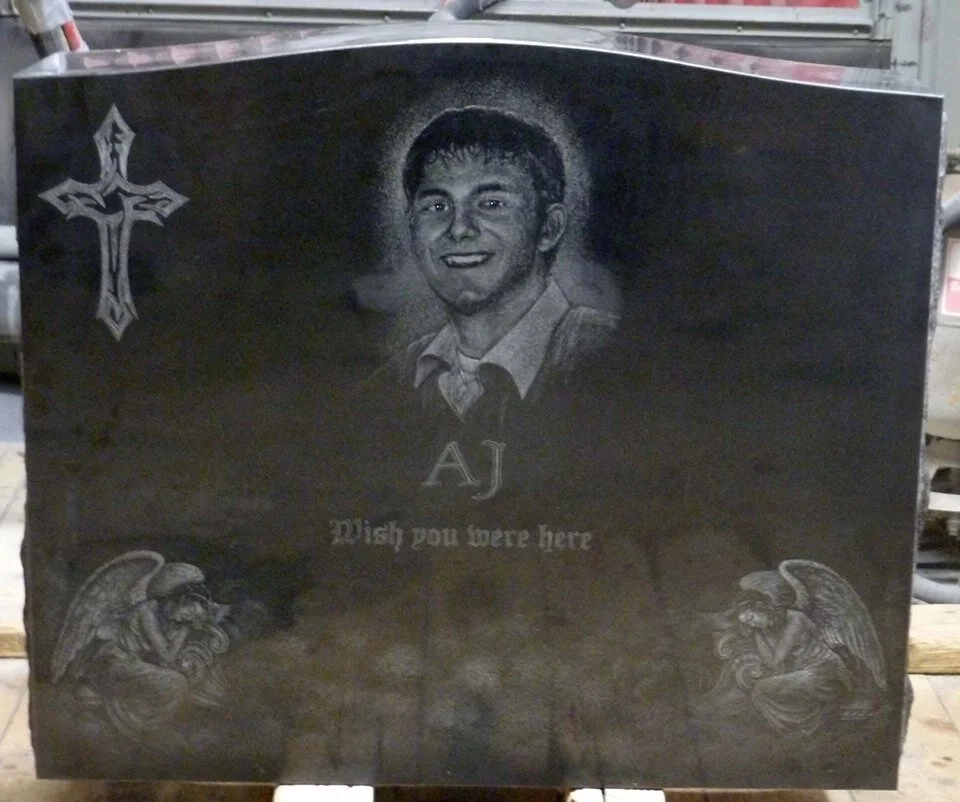 Black granite headstone with etched portrait of a young man labeled 'AJ,' featuring a cross and two angels. Inscription reads 'Wish you were here.'
