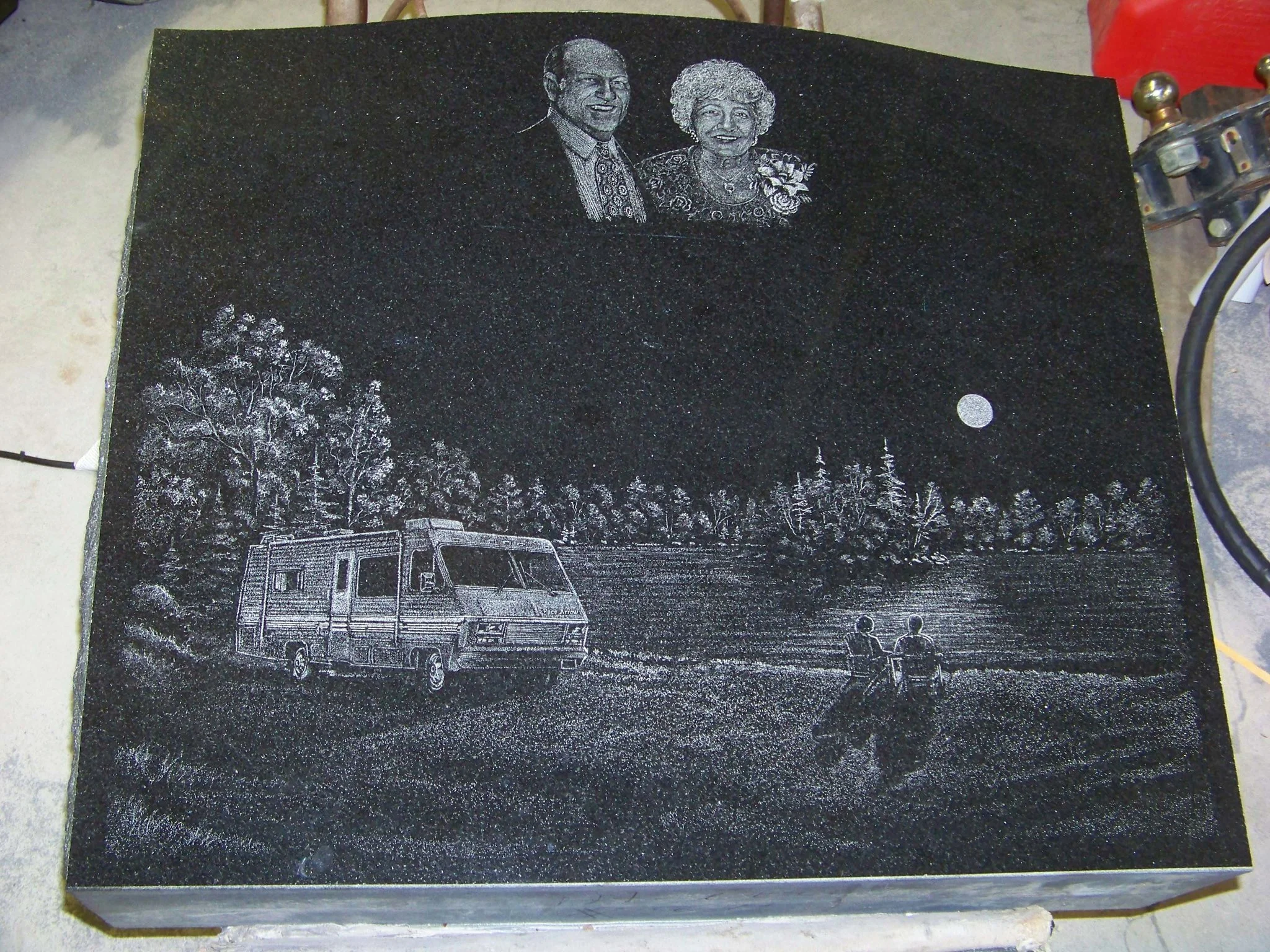 Engraved black stone with portraits of a man and woman at the top, depicting a scenic landscape with trees, a motorhome, and two people sitting by a lake under the moonlight.