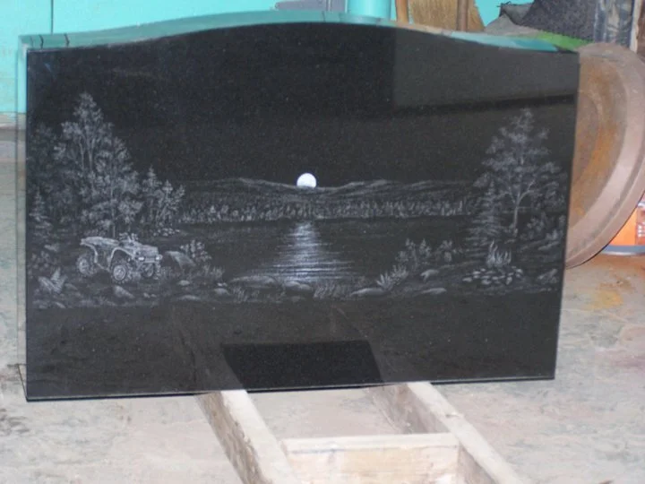 Engraved black stone with a night landscape featuring a car, trees, and a moonlit lake.