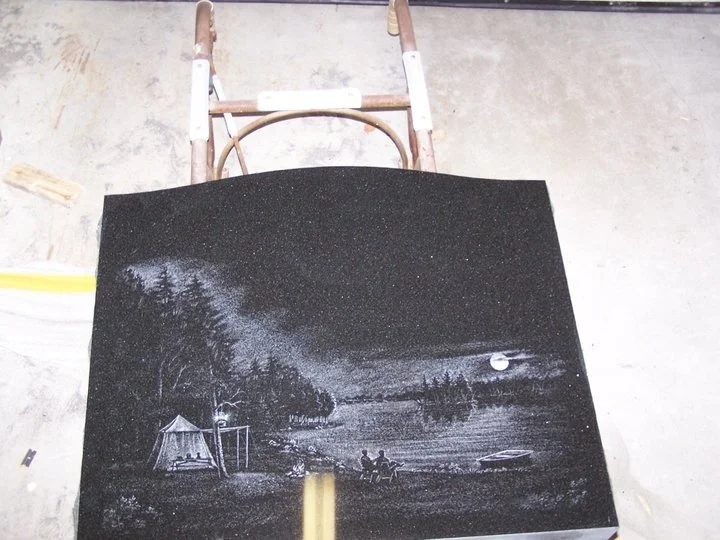 Carved black stone depicting a night scene by a lake, featuring trees, a tent, and silhouettes of people sitting. A moonlight view with a canoe on the shore is also visible.