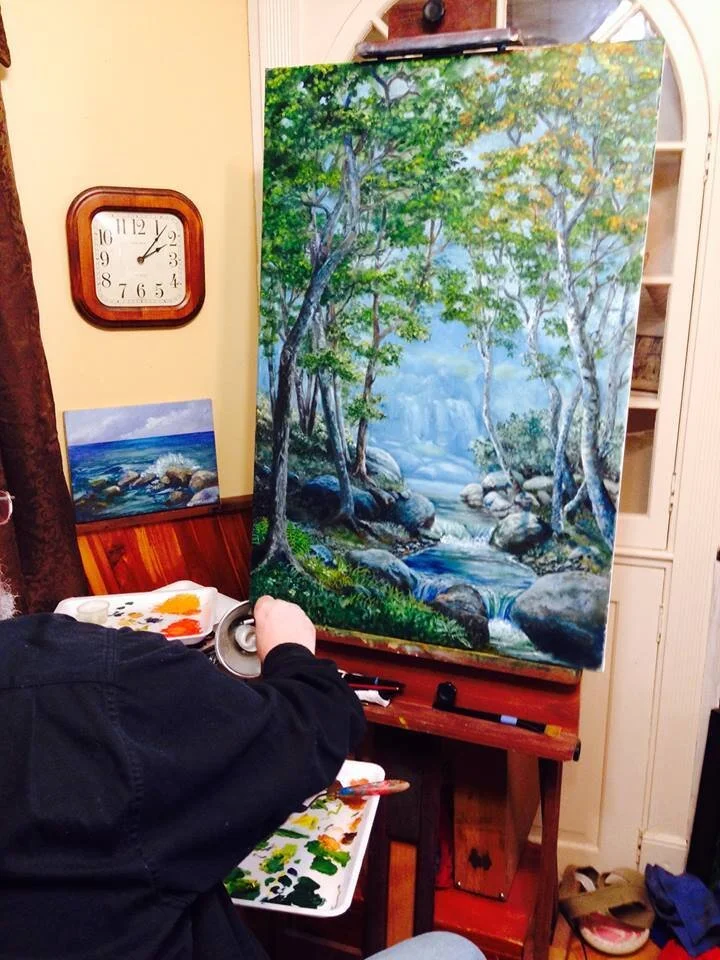 Artist painting a landscape with trees, rocks, and water on a canvas in a studio.