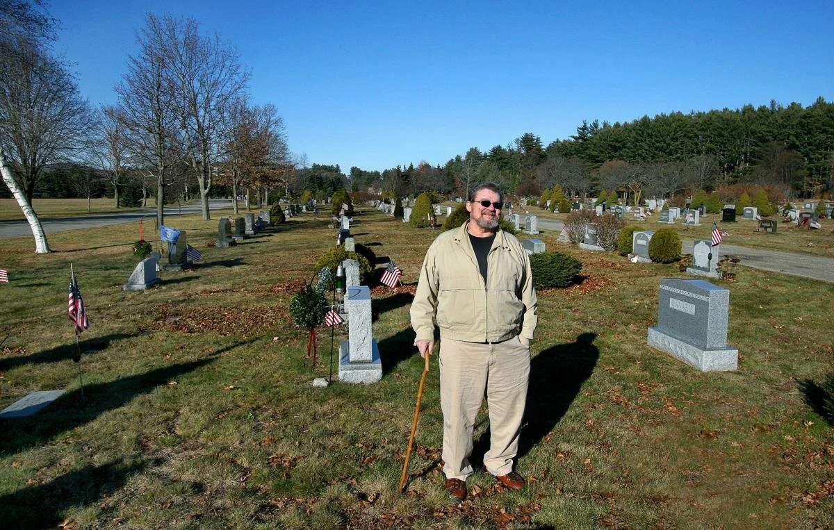 Man standing with a cane in a cemetery field, surrounded by gravestones and American flags, on a sunny day.