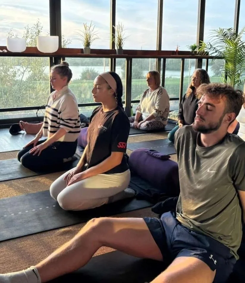 group breathwork class with men and women sitting quietly on mats with their eyes shut in a calming, light-filled space overlooking the wetlands