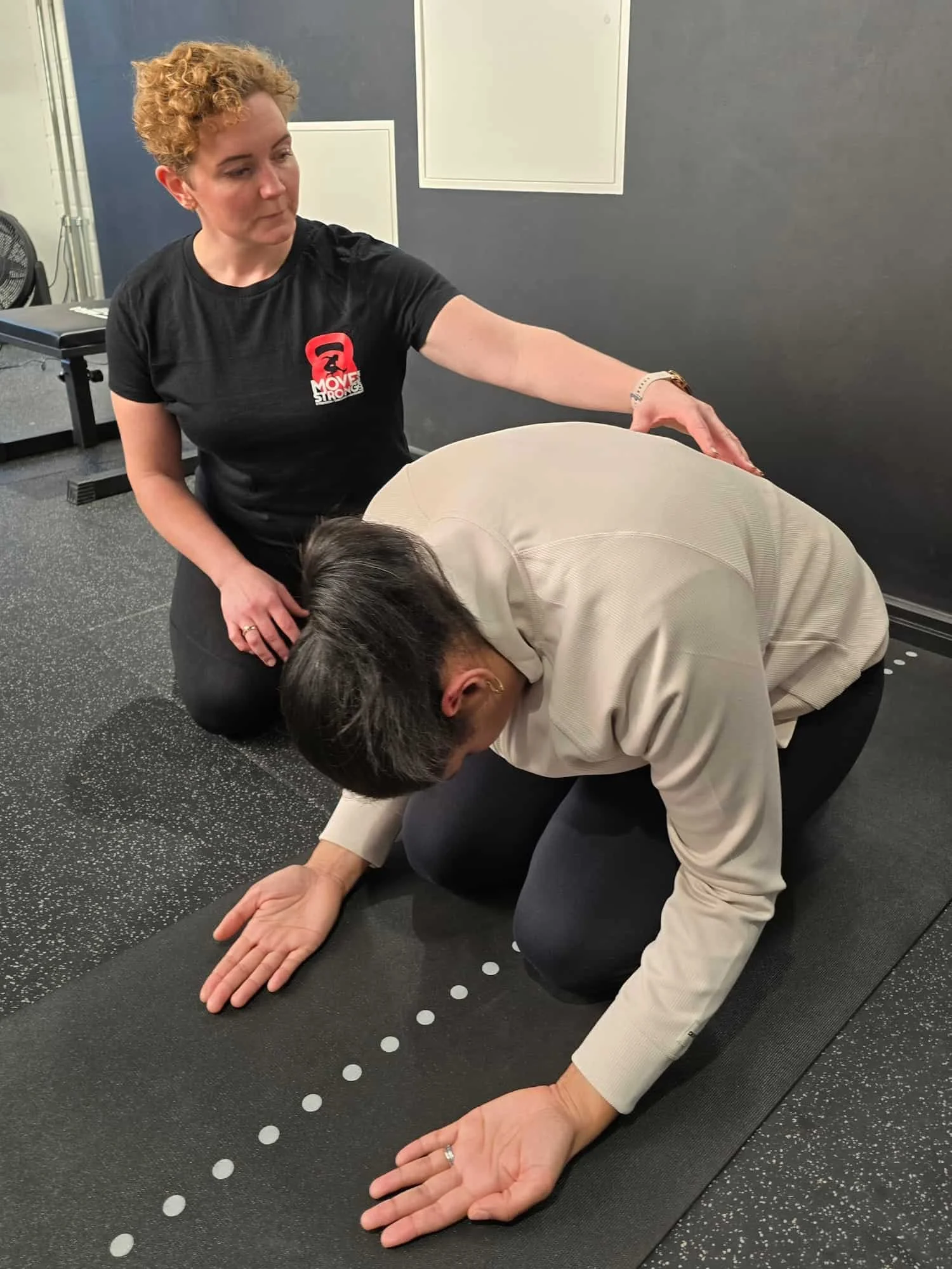 Woman on knees with head down and palms facing up, doing breathing exercises. A personal trainer is seated next to her and gently resting her hand on the woman's back as she guides her through the breathwork.