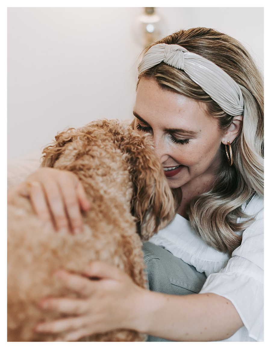 A woman with blonde wavy hair, wearing earrings and a headband, smiling and hugging a curly-haired dog.