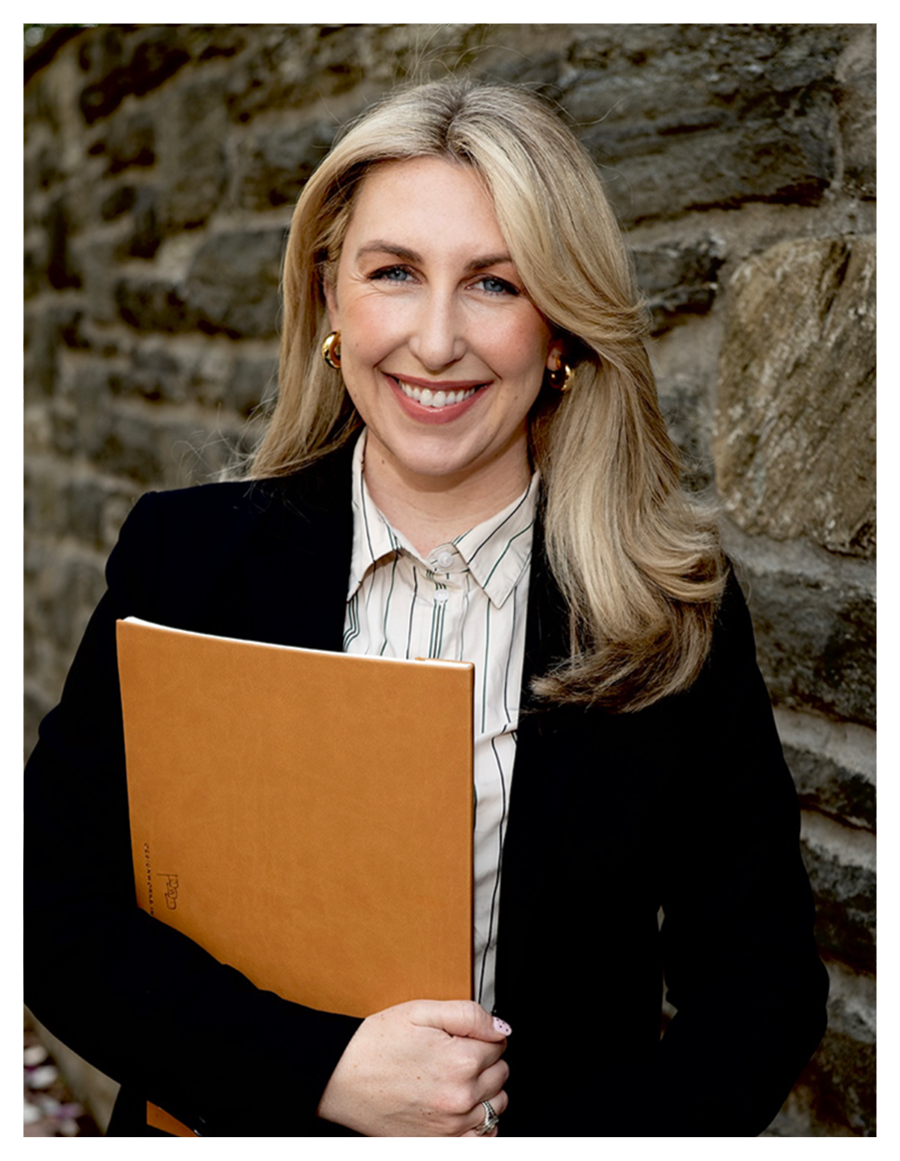 A smiling woman with blonde hair holding an orange folder, standing outdoors in front of a stone wall, dressed in business attire.
