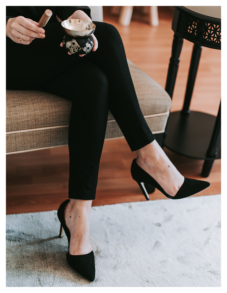Person sitting on a beige couch, wearing black high heels, black pants, and holding a tuning fork and singing bowl.