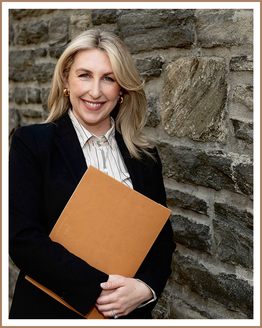 A blonde woman smiling and holding a tan folder, standing against a stone wall.