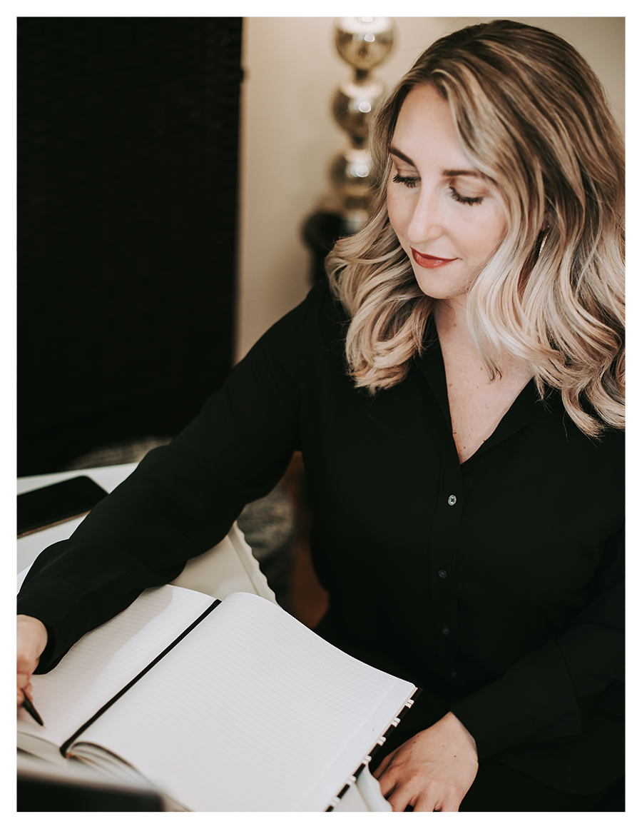 A woman with blonde wavy hair in a black shirt, sitting at a desk with an open notebook and pen, smiling and looking down.