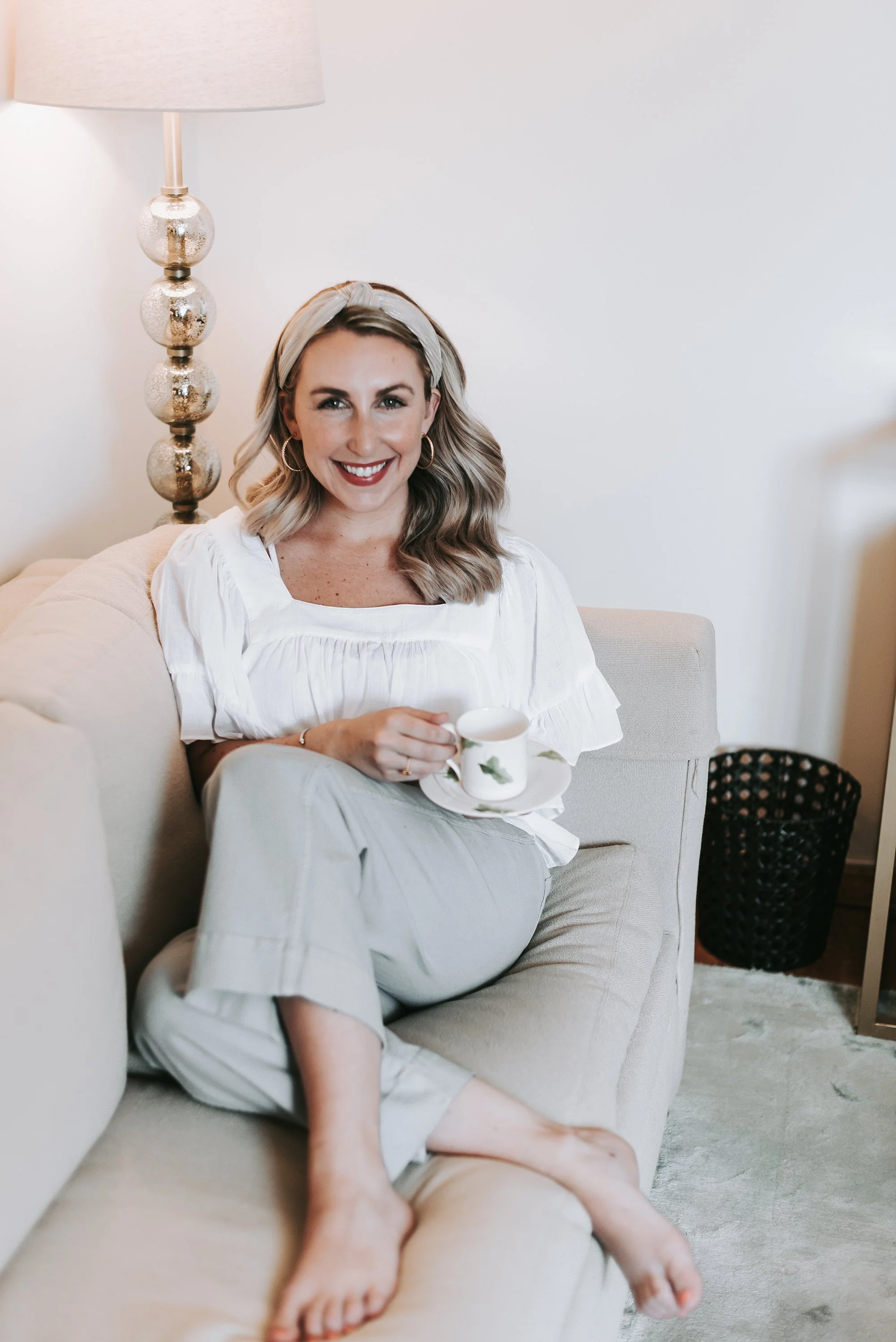 A woman with blonde hair, wearing hoop earrings and a white headband, is sitting on a cream-colored sofa, smiling, holding a mug, with her legs crossed and feet resting on the sofa, in a cozy room with a tall lamp and decorative black basket in the background.