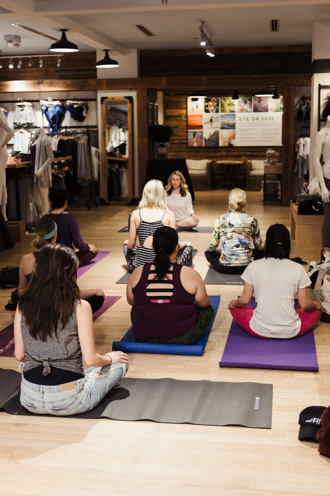 People practicing yoga in a yoga studio, sitting on mats in a cross-legged position, with a woman meditating at the front.