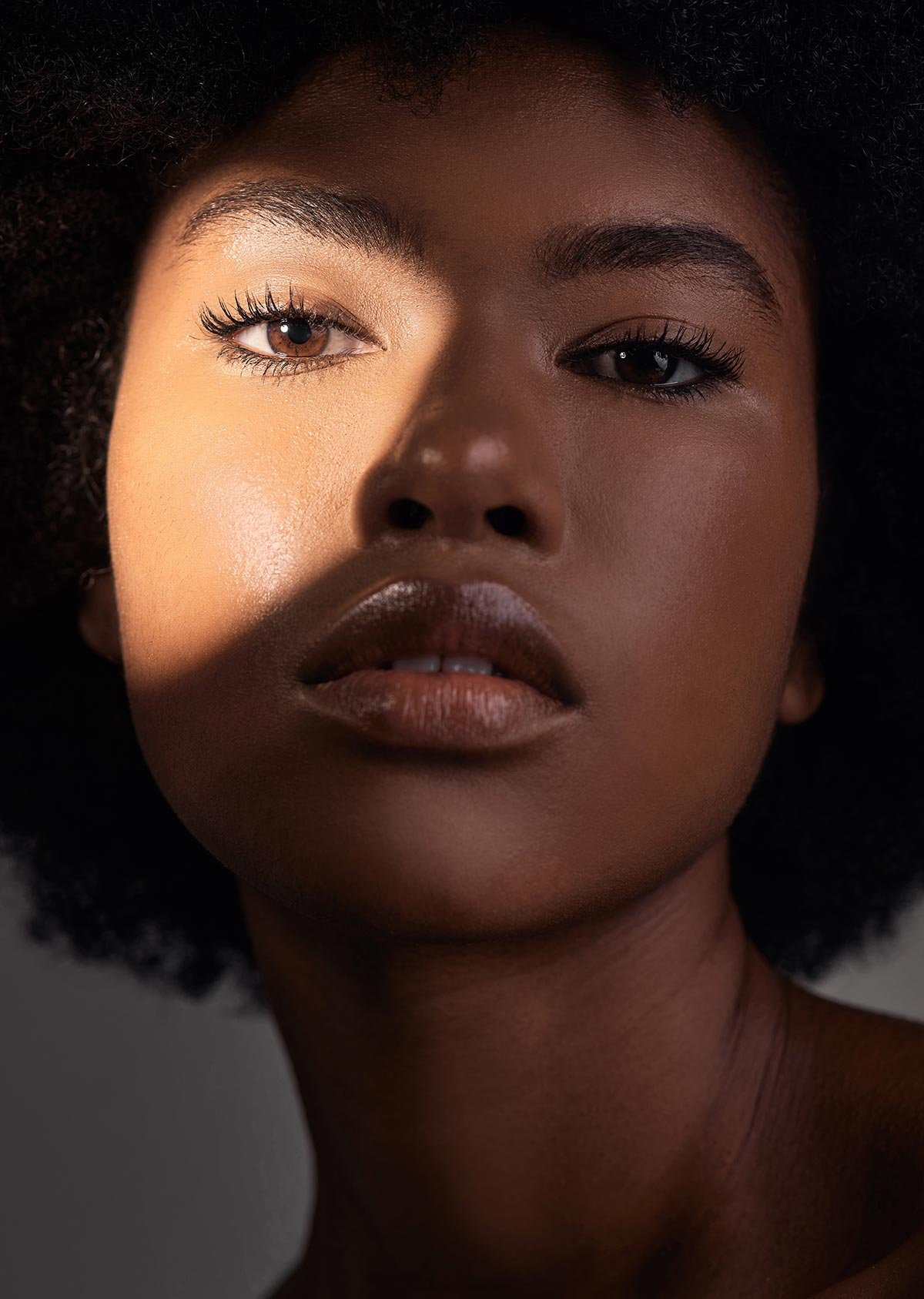 Close-up of a woman with dark skin, full lips, and natural curly hair, looking directly at the camera with dramatic lighting.