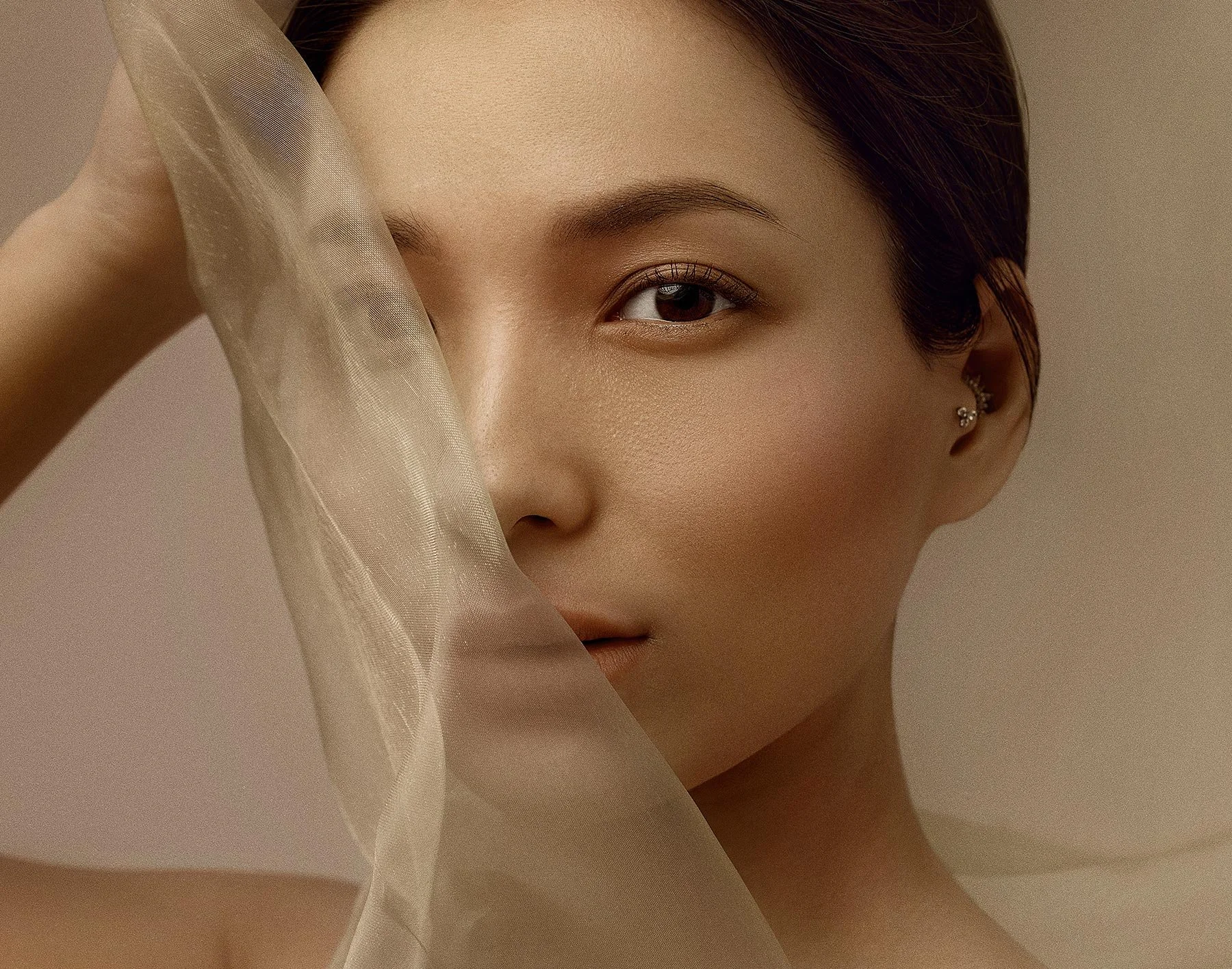 Close-up portrait of a woman with smooth skin, dark hair, and brown eyes, partially covered by sheer fabric near her face.