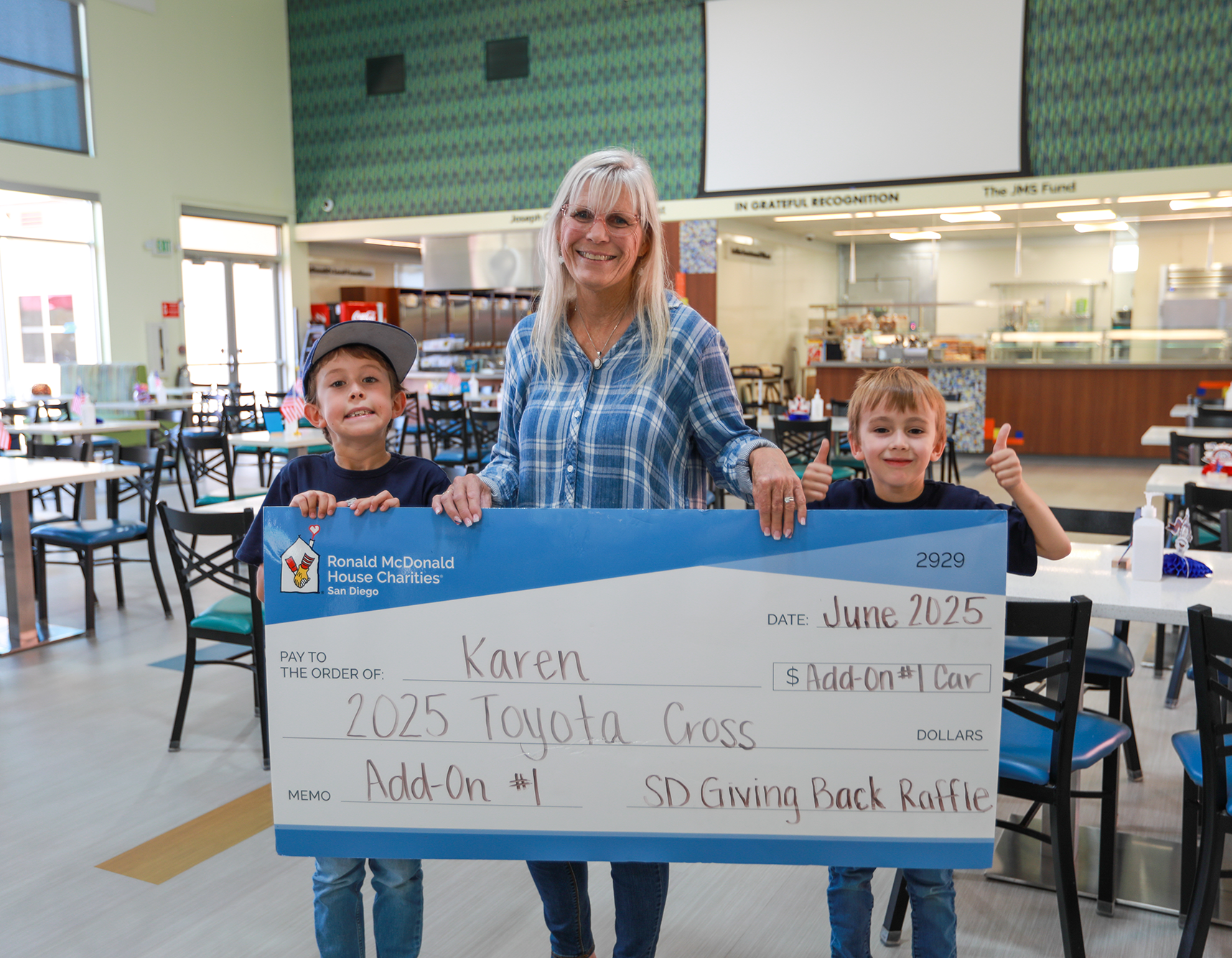 A woman and two children standing behind a large check in a cafeteria. The check is made out to Karen for a 2025 Toyota Cross, with a memo for Add-On #1, and is part of a charity raffle. The woman is smiling and the children are giving thumbs up.