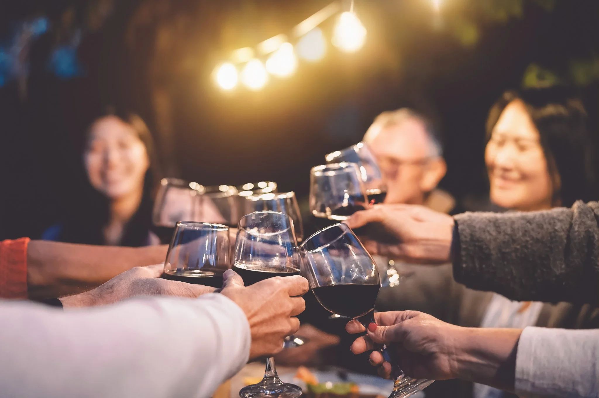 People clinking glasses of red wine in a toast at a dinner gathering, with blurred smiling faces in the background and warm overhead lighting.