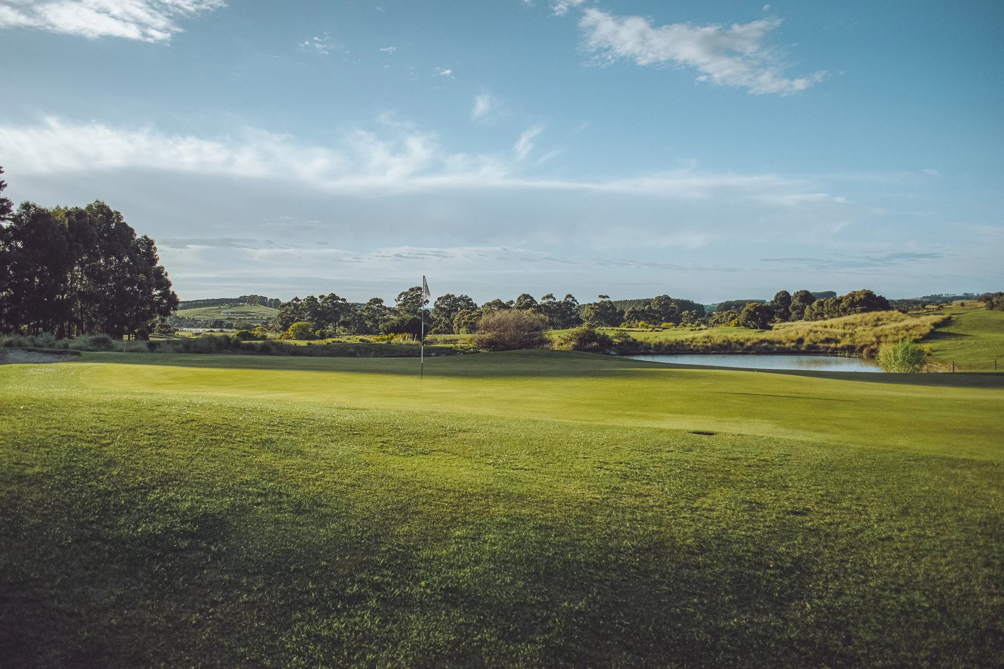 A golf course with a flag on the green, water features in the background, and trees under a partly cloudy sky.