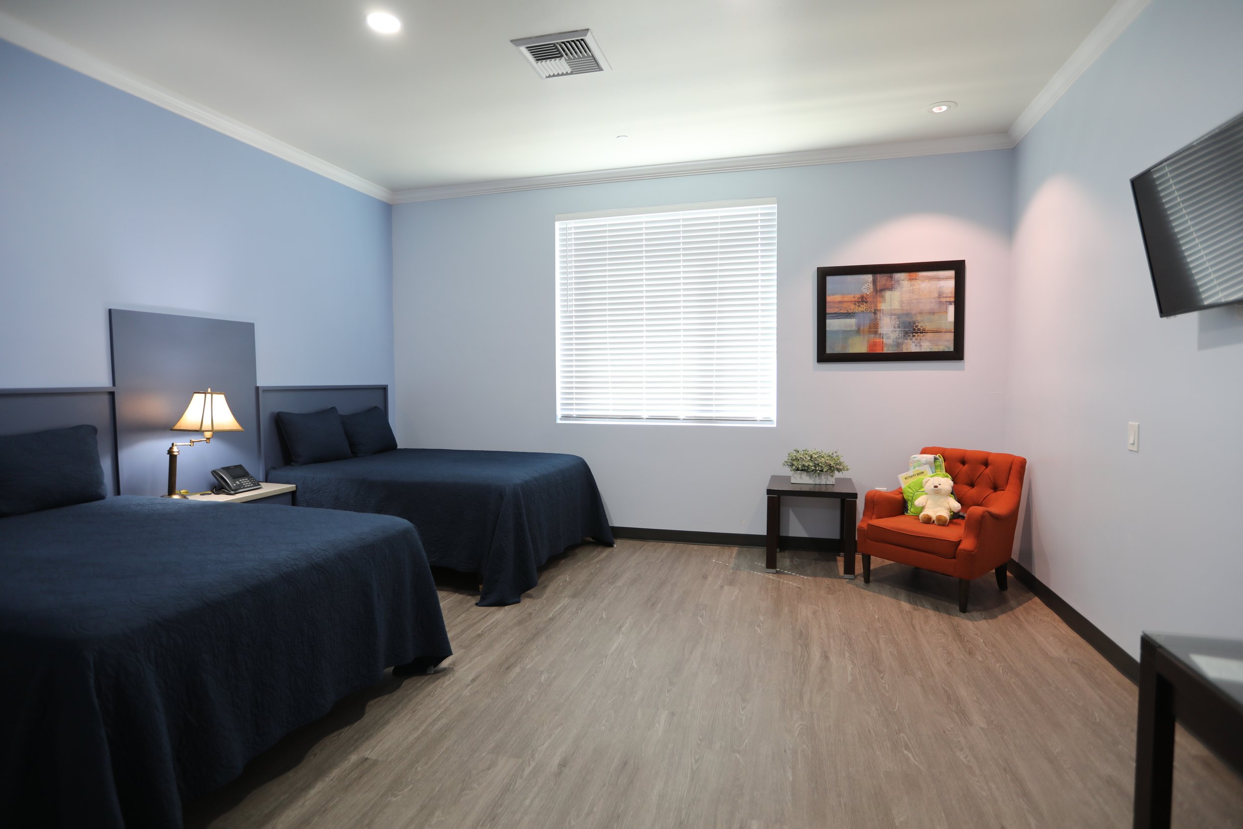 Hotel room with two beds with dark blue bedding, a nightstand with a lamp and phone, a window with blinds, a red armchair with a teddy bear, a small side table, a wall-mounted TV, and a framed abstract art piece.