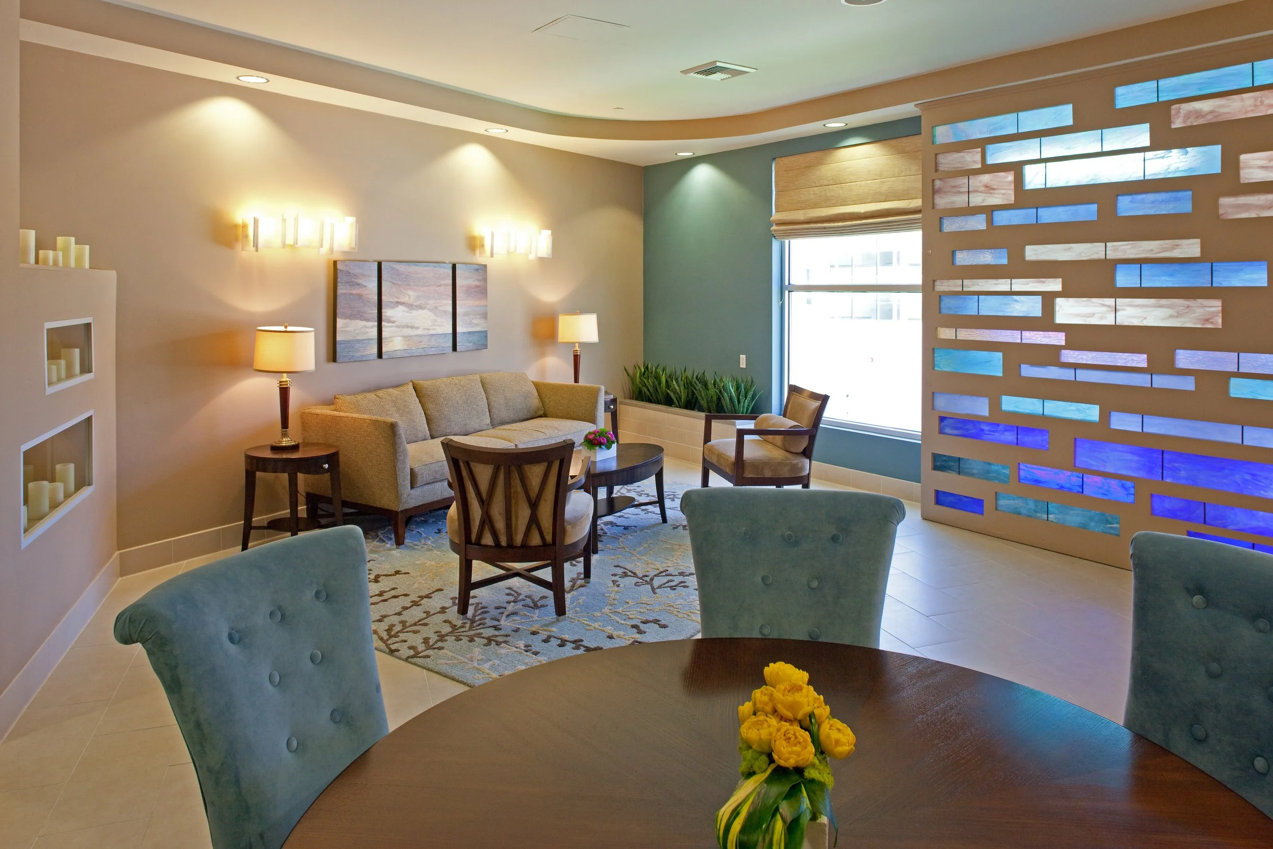 A cozy hotel lobby with a seating area, including a beige sofa, chairs, and a coffee table with flowers. Soft lighting, artwork on the wall, and a window with blinds are also visible.