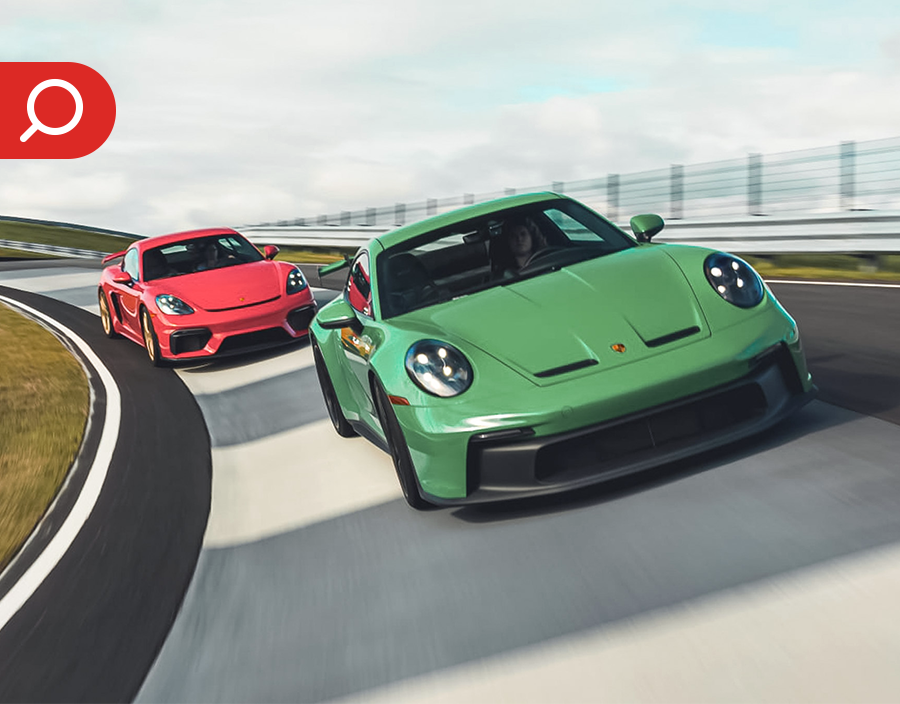 Two sports cars, a green Porsche and a red Porsche, racing on a track with curve and fencing in the background.