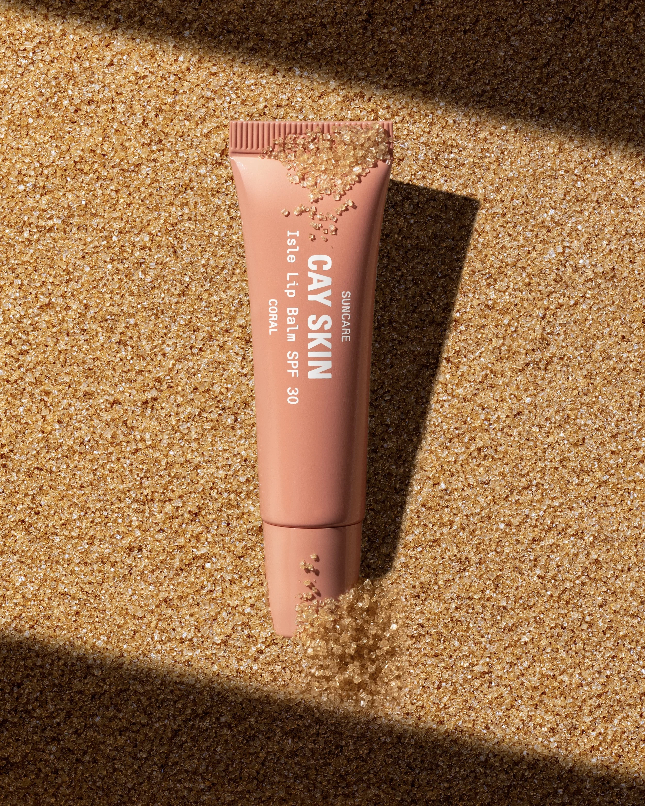 A tube of CAY SKIN sunscreen lip balm labeled 'Suncurse,' with a coral color tube, partially buried in sand, casting a shadow.
