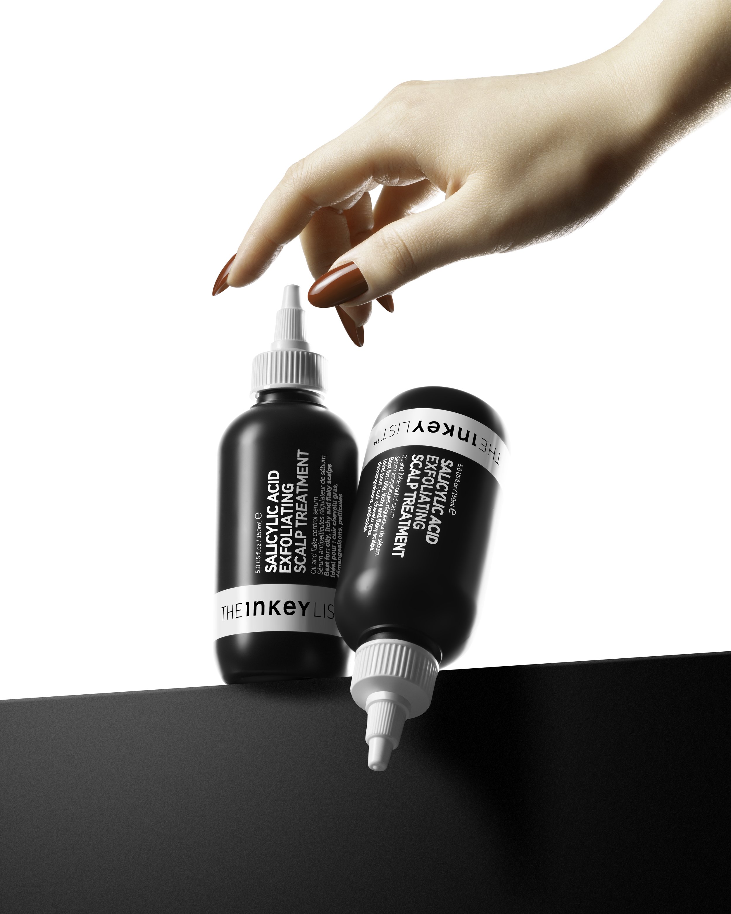 A woman with red painted nails suspends two black bottles of salicylic acid exfoliating scalp treatment, one standing upright and the other lying down, against a plain white background and black surface.