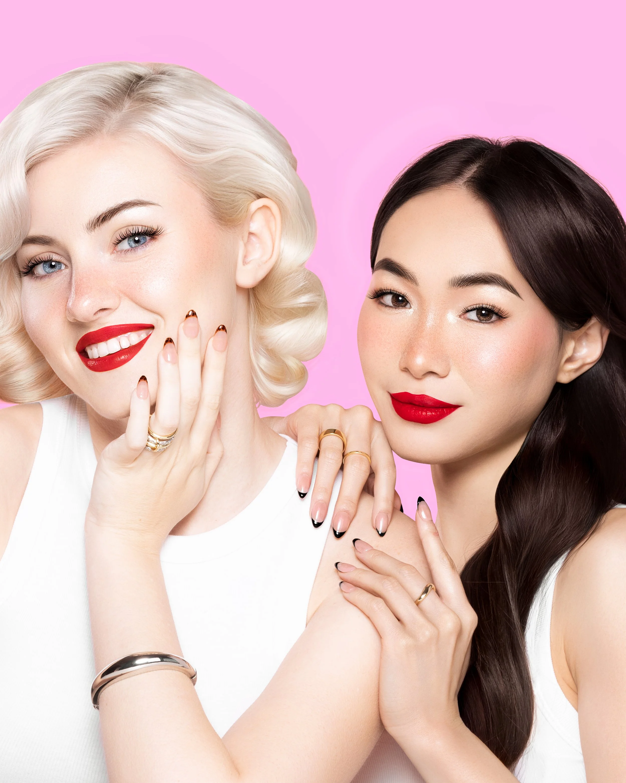 Two women with makeup and jewelry, smiling, against a pink background.