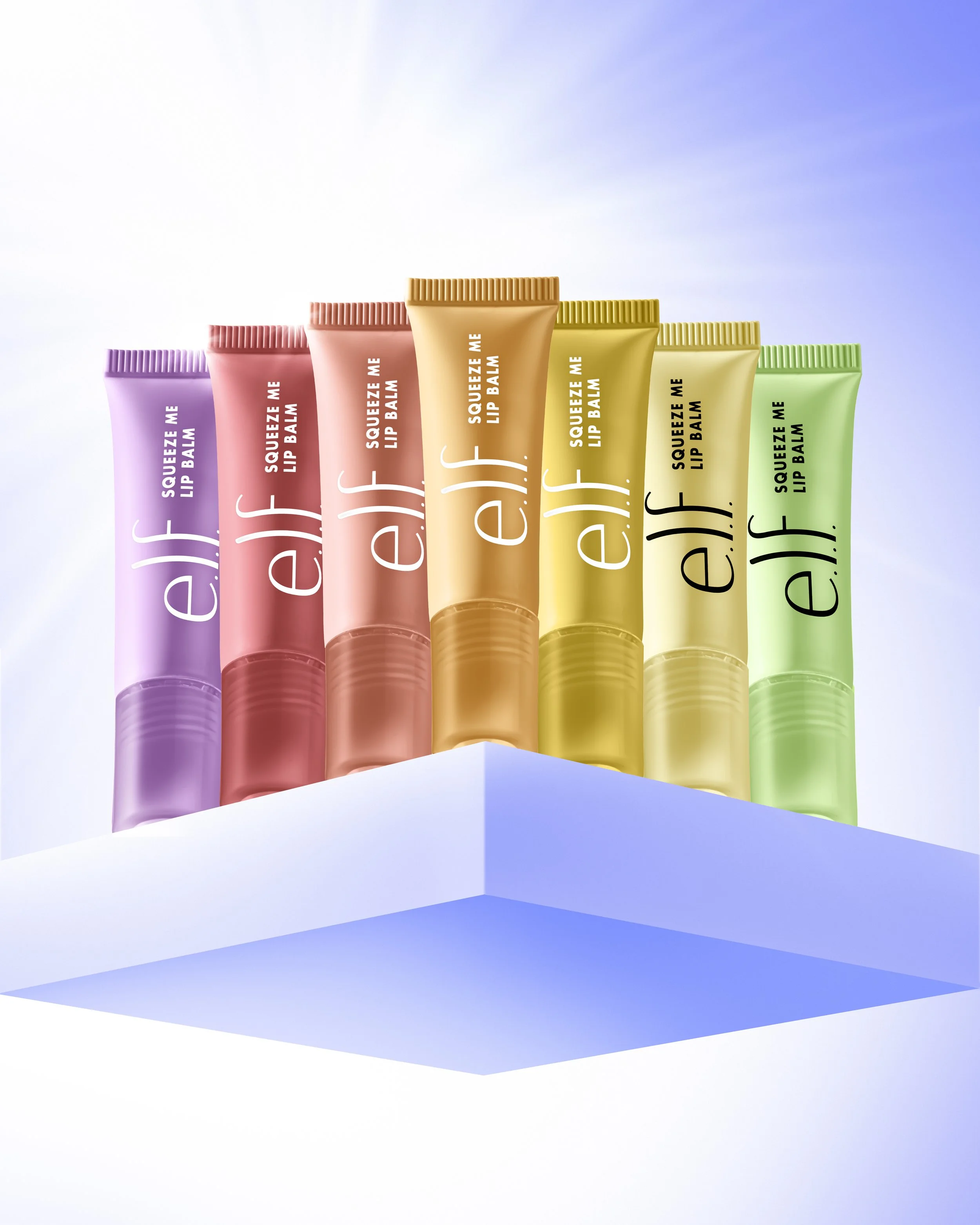 Six colorful tubes of e.l.f. lip balm arranged on a white platform with a blue sky background.
