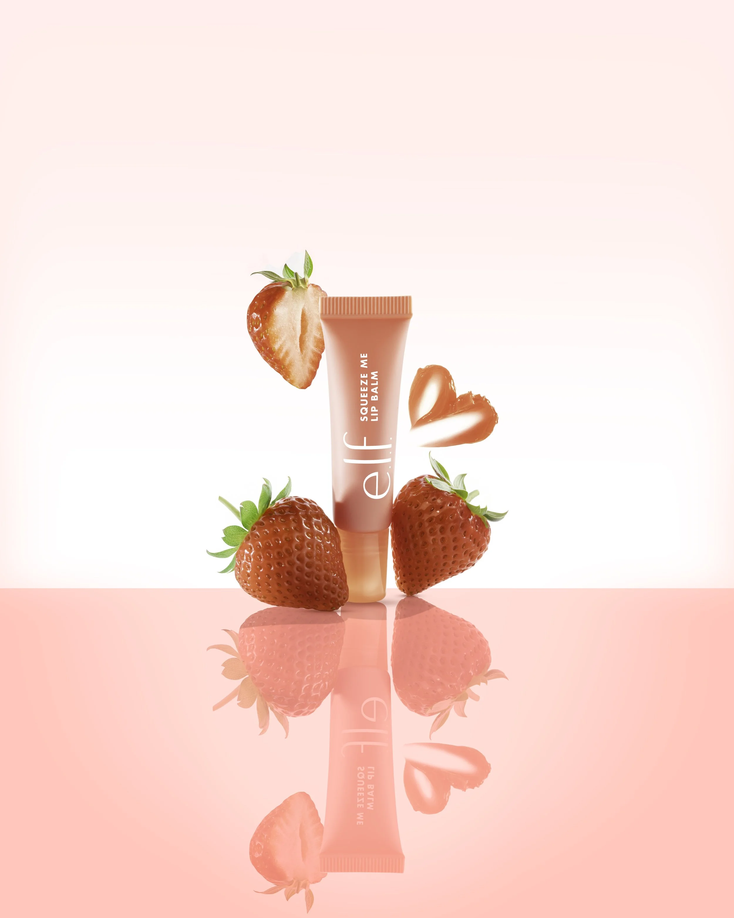 A pink tube of elf Squeeze Me Lip Balm surrounded by fresh strawberries and strawberry-shaped lip balm shapes, reflected on a shiny pink surface.