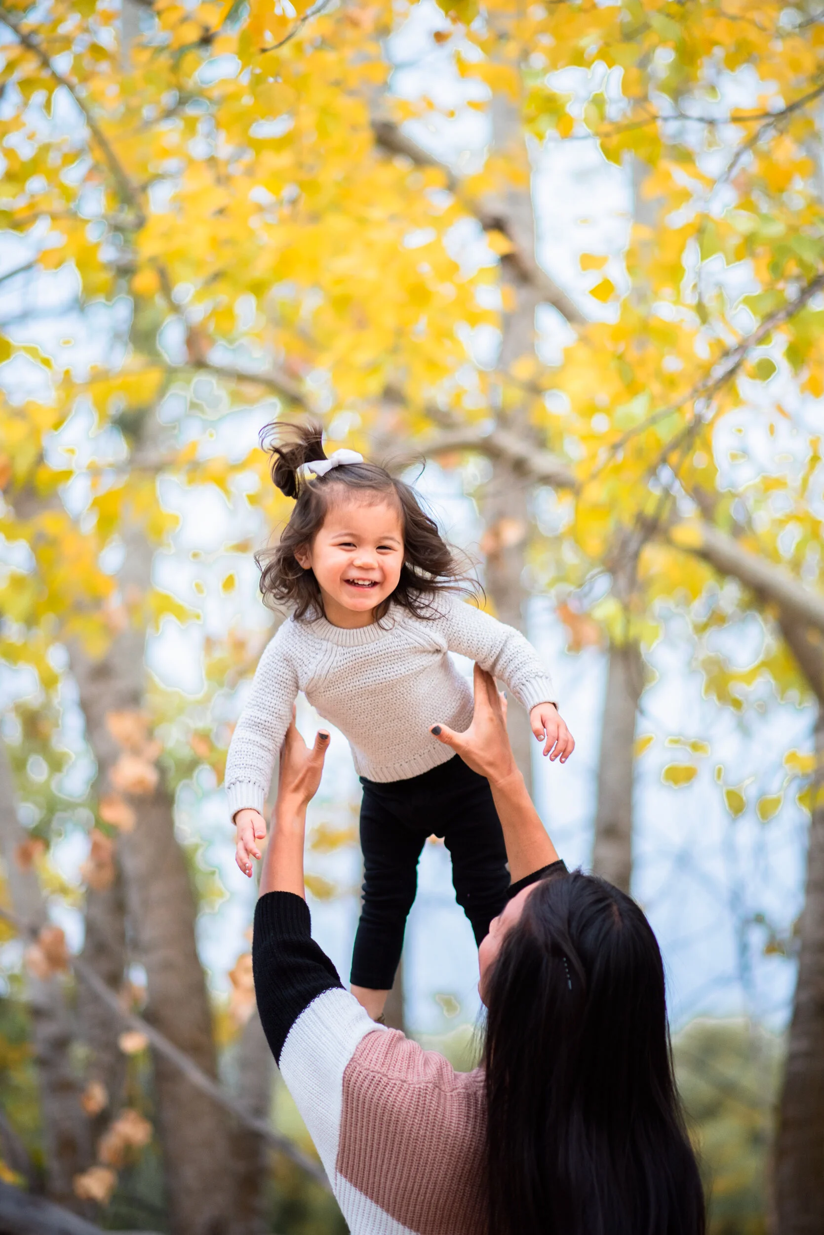 A young girl smiling as she is lifted in the air by an adult, with her arms outstretched, surrounded by yellow autumn leaves on trees.
