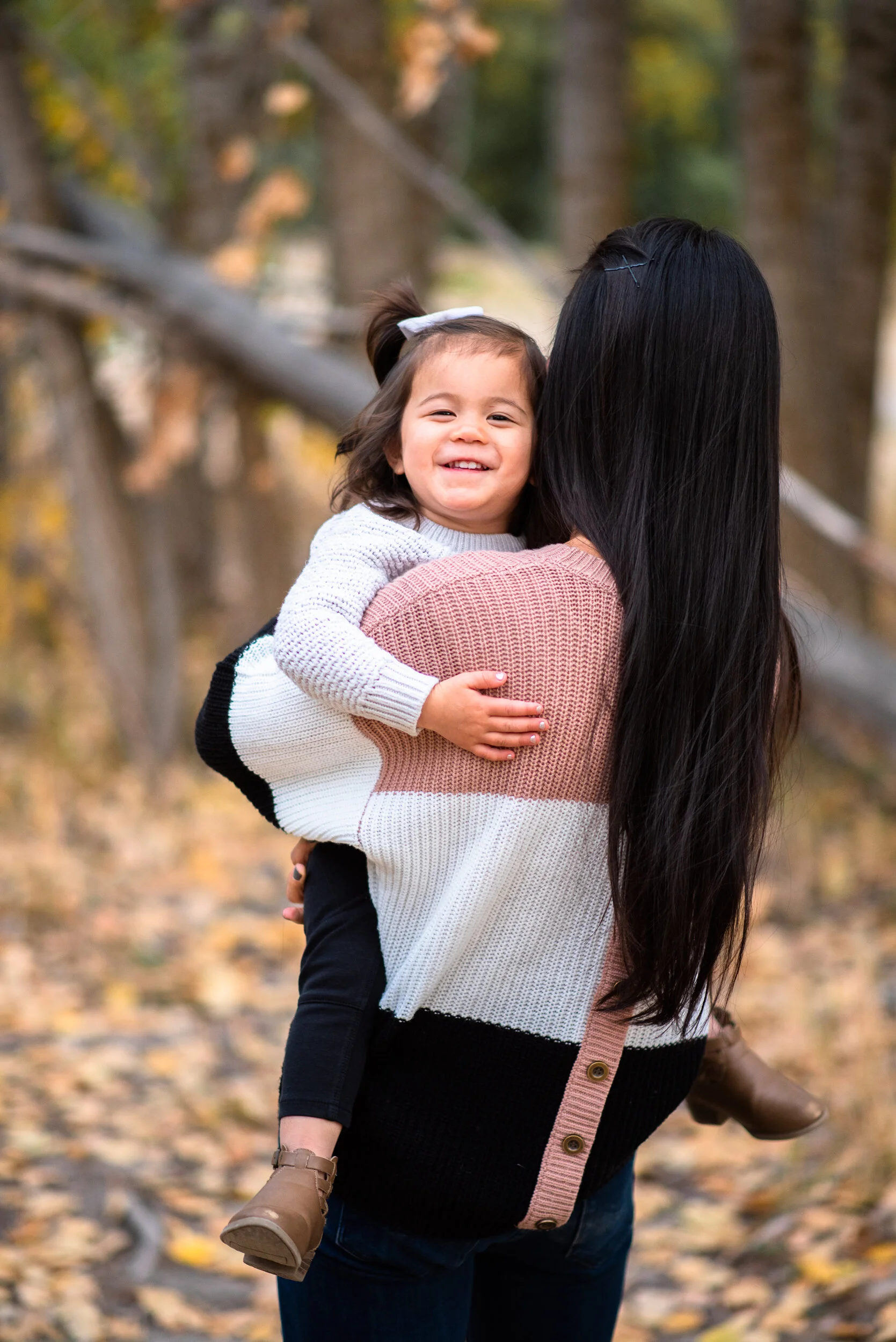 A woman holding a smiling young girl in an outdoor wooded area during autumn.